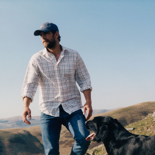 A man in a plaid shirt and cap walks with a black dog on a sunny hillside. The landscape features rolling hills under a clear blue sky, evoking tranquillity.