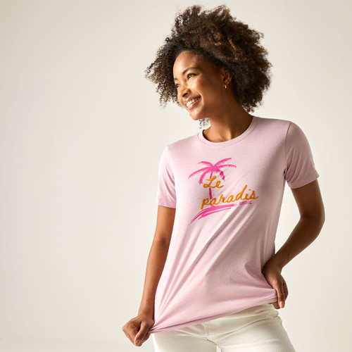 Woman wearing a light pink short-sleeve T-shirt with a palm tree graphic, smiling and holding the hem of the top.