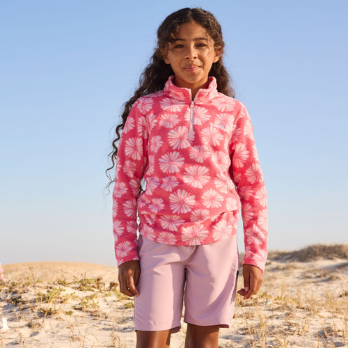 Young girl in a pink floral jumper and light pink shorts stands on a sandy beach, smiling against a clear blue sky. The scene is bright and cheerful.