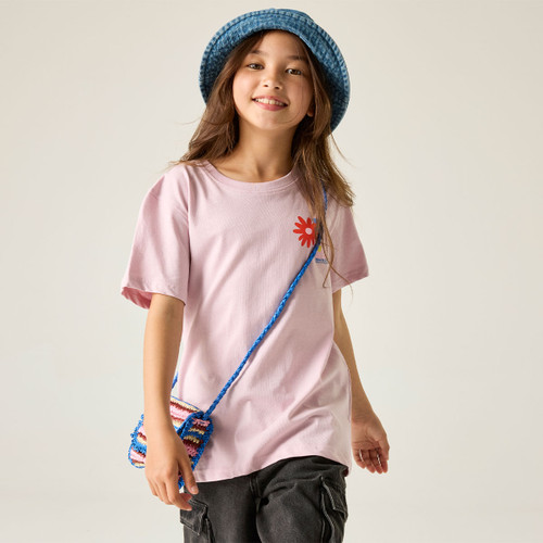 A smiling child wears a pink t-shirt with a red flower, a blue hat, and a colourful knitted purse. The background is plain, conveying a playful tone.