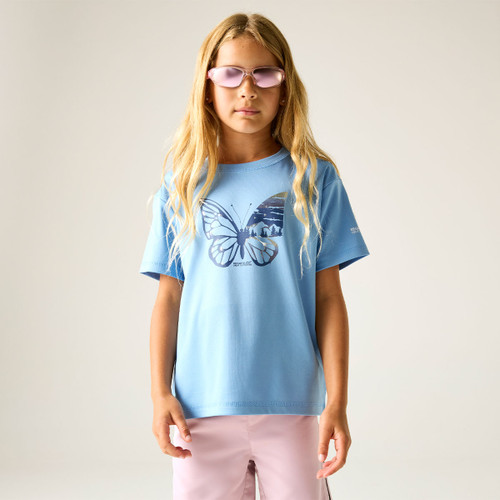 A young girl with long blonde hair wearing pink sunglasses, a light blue butterfly-print t-shirt, and pink pants, standing against a plain backdrop.