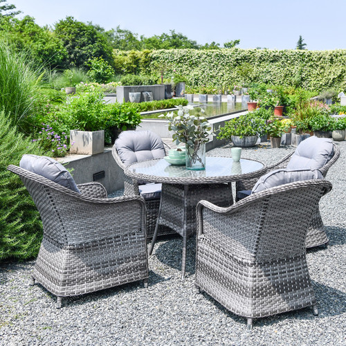 Grey rattan round dining table with four cushioned armchairs set on a gravel patio, styled with tableware in a landscaped garden.
