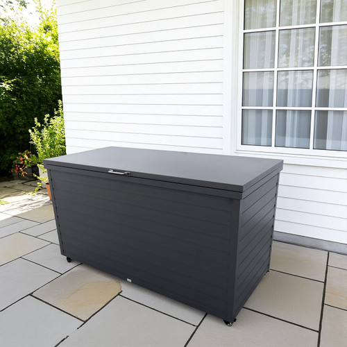 Large grey storage box on patio with grey tiles, against a white house exterior. A window with white frames is on the right. Lush greenery in the background.