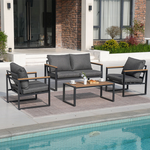 Modern outdoor patio with dark gray cushioned seating and wooden coffee table. Set on a rug beside a pool, brick house backdrop, and greenery.