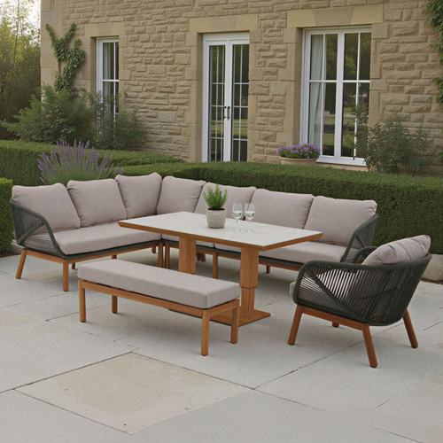 Outdoor corner dining set with an L-shaped sofa and armchair in dark fern green rope weave and grey cushions, a rectangular table with a pale top and wooden base, and a matching bench on a stone patio.