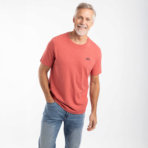 Man wearing a coral-red Weird Fish T-shirt and blue jeans, smiling and leaning slightly forward against a white studio background.