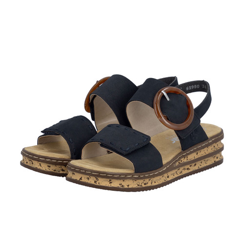 Pair of blue Rieker sandals with wide crossover straps, touch-fastening front strap and large tortoiseshell-effect buckle, on a tan footbed with a speckled wedge sole, shown at a three-quarter angle on a white background.