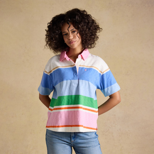 Model wearing a Joules women’s short-sleeve rugby polo top with bold blue, green, white and pink stripes and a pink collar, styled with blue jeans.