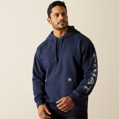 Three-quarter front view of a model wearing a navy pullover hoodie with a kangaroo pocket and large “ARIAT” logo down the sleeve.