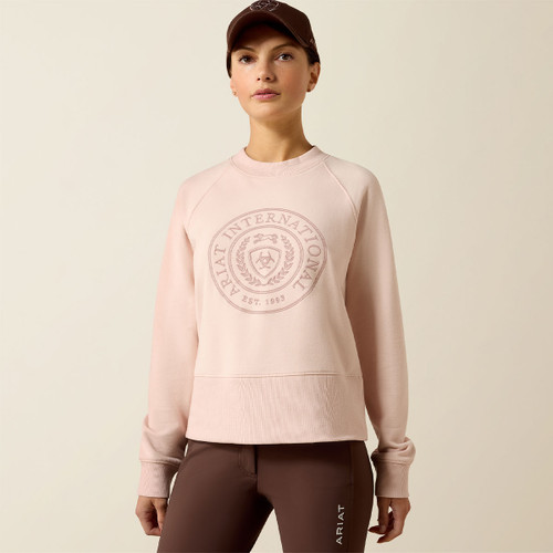 A person in a light pink sweatshirt with "Ariat International" logo stands against a neutral background. They wear a brown cap and brown pants, looking forward.