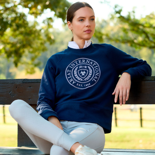 A person in a navy blue sweatshirt with white lettering and a logo sits relaxed on a bench in a sunny park. The scene conveys tranquility and style.
