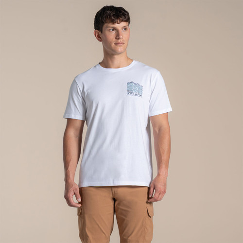 Male model wearing a white Craghoppers T-shirt with small blue mountain chest print, paired with tan trousers, standing against a beige backdrop.