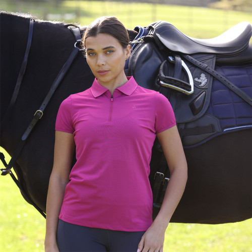 Rider wearing a bright pink short-sleeve zip-neck polo shirt, standing beside a saddled horse outdoors.