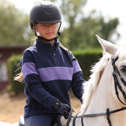 Young rider wearing a navy and lilac Aubrion Equestrian colour-block fleece riding a white horse in an outdoor arena.