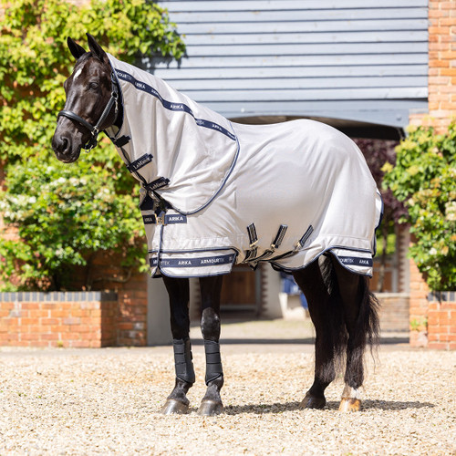 A black horse wearing a light grey protective sheet stands on a gravel path in front of a brick building with green foliage. The setting is calm and sunny.