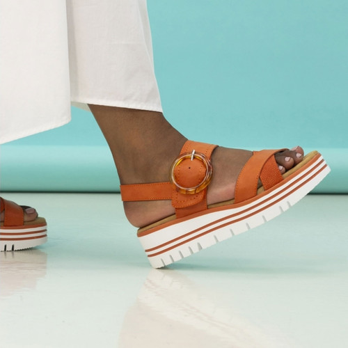 Close-up side view of orange platform sandals on a model’s feet, featuring a large round buckle and striped white sole against a blue backdrop.