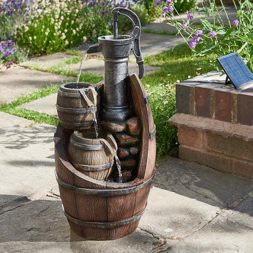 Rustic garden water feature with a hand pump pouring water into stacked wooden barrels, set on a stone patio with surrounding flowers.