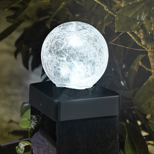 Close-up of a crackle-glass ball garden light glowing white, mounted on a dark fence post surrounded by foliage at night.
