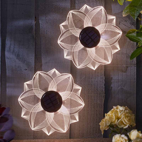 Set of two solar-powered flower-shaped wall lights with intricate line patterns, glowing warm white on a wooden fence at night.