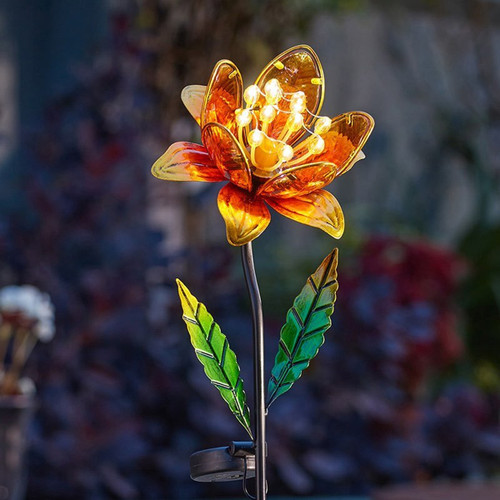 Solar-powered flower garden stake light with amber and yellow petals and glowing LED centre, standing among plants at night.