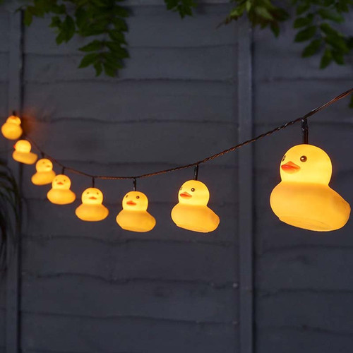 String of glowing yellow rubber duck fairy lights hanging outdoors against a garden fence at night.