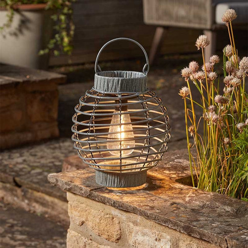 Slate-grey wire cage lantern with a glowing bulb inside, sitting on a stone ledge beside garden plants.