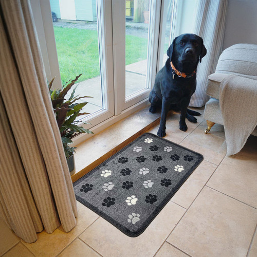 A black dog wearing an orange collar sits by a glass door on a tiled floor. Beside it is a grey mat with black and white paw print designs.