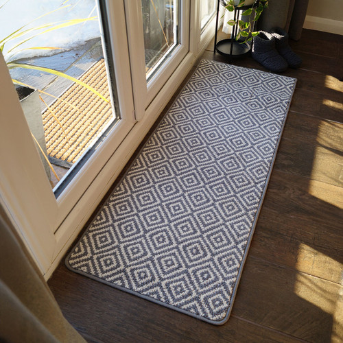 Grey and white diamond-patterned runner rug on wooden floor beside glass doors. Sunlight streams in, casting shadows. Cosy slippers and a plant nearby.