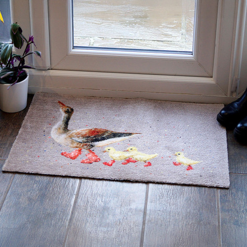 A charming doormat with a painted goose leading three ducklings is placed by a glass door. A potted plant and black boots are nearby on the wooden floor.