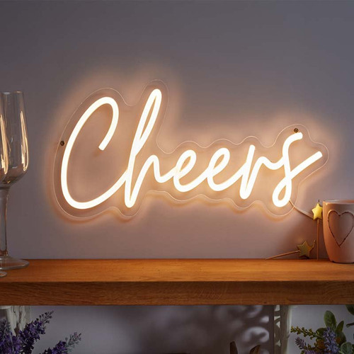 Warm white neon “Cheers” sign in cursive on a wall above a wooden shelf, with a tall champagne flute on the left and a mug on the right.