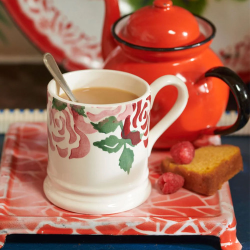 Half-pint mug with pink and red rose design, filled with tea on a patterned tray beside a red teapot, spoon and slice of cake.