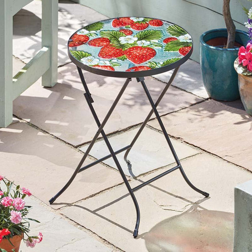Round folding side table with a strawberry-patterned top on a patio beside potted plants.