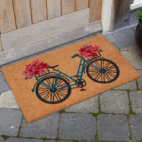 Rectangular coir doormat with a bicycle and flower basket design, placed outside a wooden door on stone paving.
