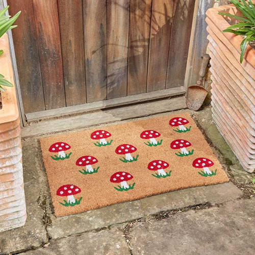 Rectangular coir doormat with a red mushroom pattern, placed outside a wooden garden gate on stone paving.