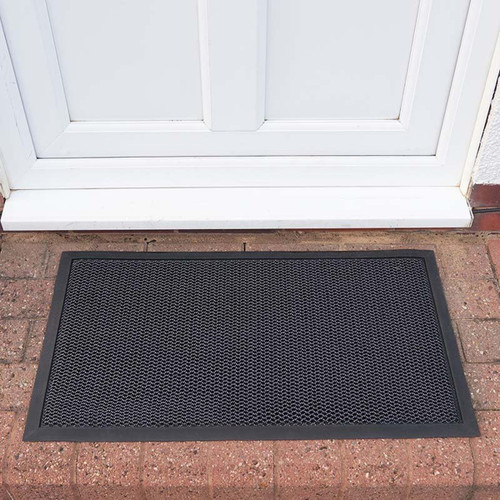 Rectangular black scraper doormat placed outside a white door on brick paving.