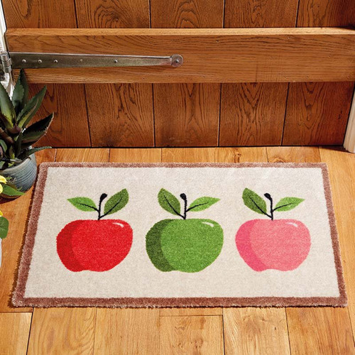 Rectangular doormat with a light background and brown border, featuring three apples (red, green, and pink) with leaves, placed on a wooden floor in front of a wooden door.
