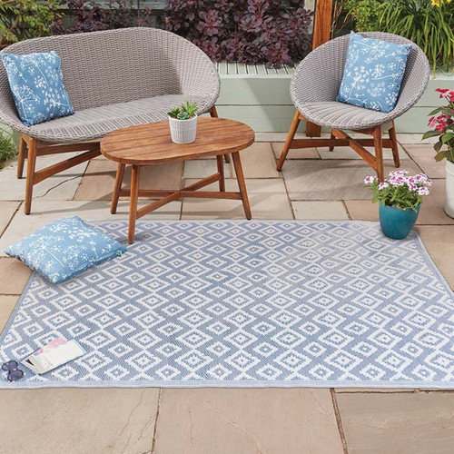 Light blue and white diamond-pattern outdoor rug on a patio, styled with wicker lounge seating, a wooden coffee table, and blue cushions.