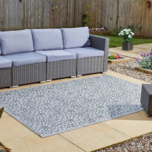 Grey wicker patio sofa with pale blue cushions beside a large grey outdoor rug featuring a white floral lattice pattern on a stone patio, with a fenced garden and flower beds in the background.