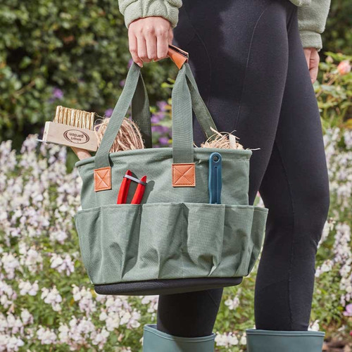 A person holding a green gardening tote with long handles and multiple pockets, filled with tools like pruners, a trowel, twine, and plant labels, in front of a flower-filled garden.