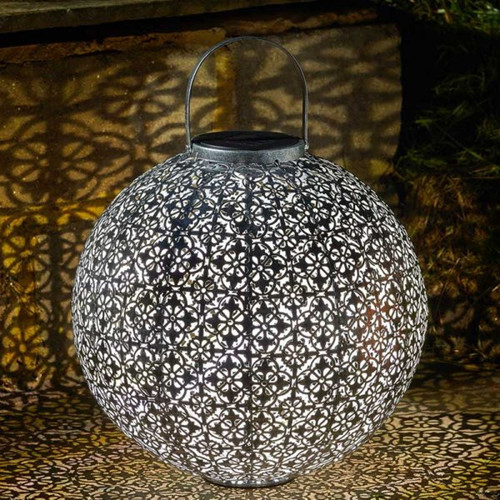Illuminated metal sphere lantern with an intricate floral cut-out pattern, casting patterned light onto the ground at night.
