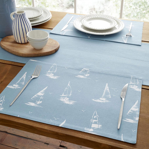 Blue cotton placemat with white sailboat print set on a wooden dining table, styled with white plates, cutlery and matching coastal table linens.