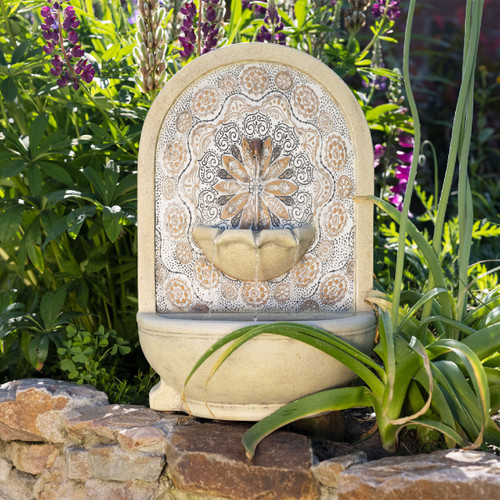 Decorative garden fountain with a floral pattern, set against a backdrop of green leaves and purple flowers, creating a serene, natural scene.