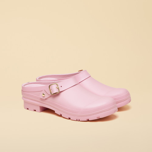 Pair of pastel pink rubber clogs with a gold buckle strap, photographed on a beige gradient background.