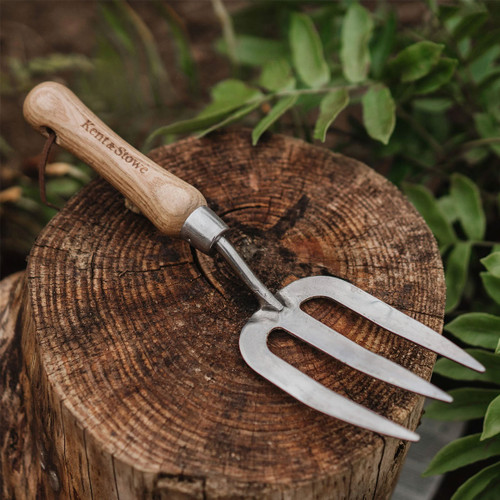 Wood-handled garden fork rests on a tree stump, surrounded by green leaves. The scene conveys a rustic, peaceful gardening atmosphere.