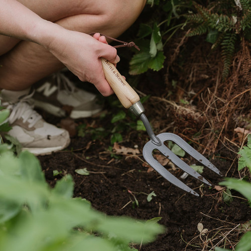 A person wearing sneakers is crouched down in a garden, using a hand fork to dig into dark soil. Green plants and leaves surround the scene, suggesting gardening activity.