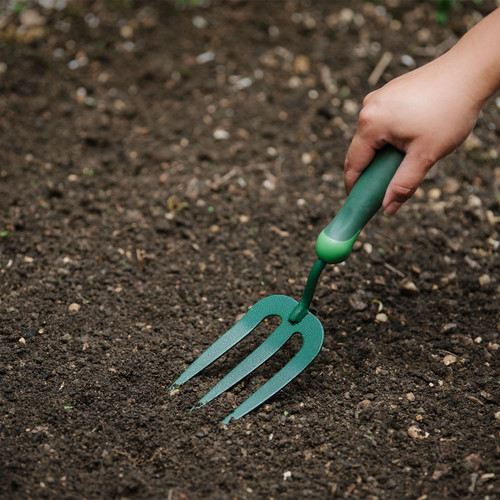 A hand holds a green gardening fork, preparing soil scattered with small pebbles, suggesting the start of a gardening activity.