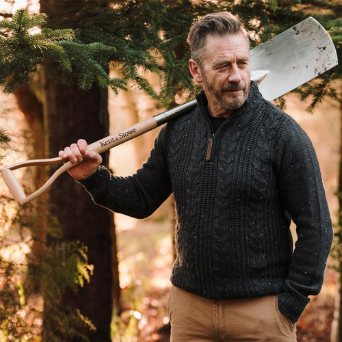 Man in a dark jumper stands in a forest, holding a shovel over his shoulder. Sunlight filters through trees, creating a warm, contemplative atmosphere.