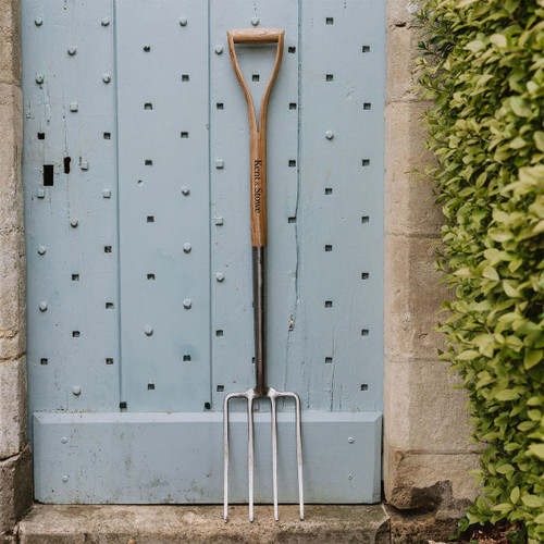 A long-handled garden fork leans against a textured light blue door with small square studs. Lush green foliage frames one side, creating a serene gardening scene.