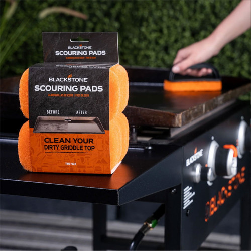 Blackstone scouring pads two-pack displayed on a flat top griddle, with an orange scrub pad handle cleaning the cooking surface in the background.