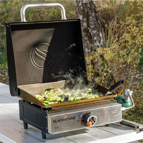 Blackstone single burner tabletop gas griddle with hood open, cooking mixed vegetables outdoors on a picnic table.
